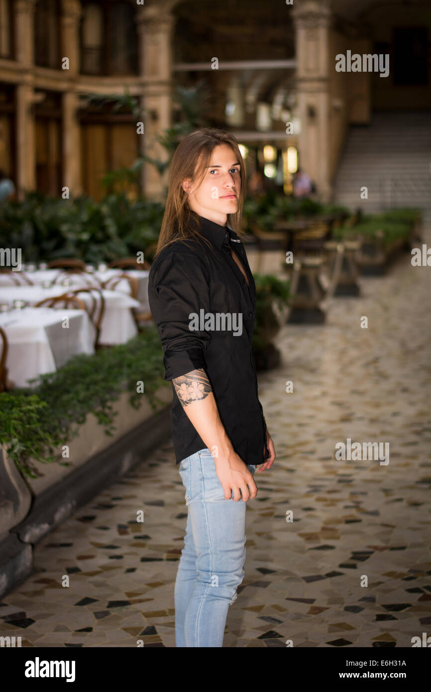 Bello capelli lunghi uomo ritratto in camicia nera in piedi in galleria elegante in città europea centro Foto Stock