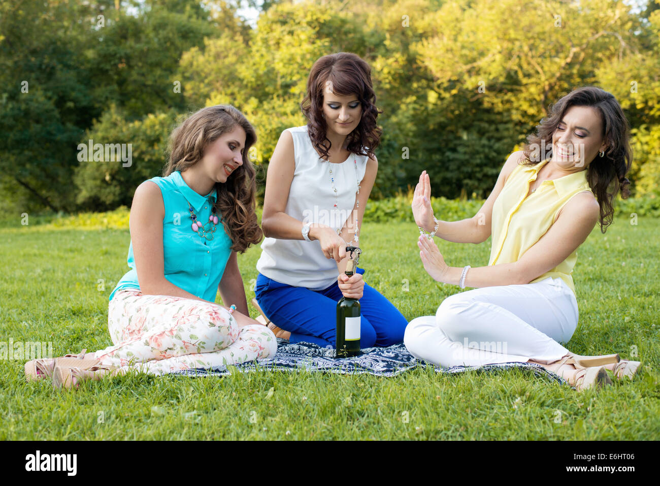Tre giovani piuttosto fidanzate apertura di una bottiglia di vino sul prato durante il picnic, ridendo e divertendosi. Foto Stock