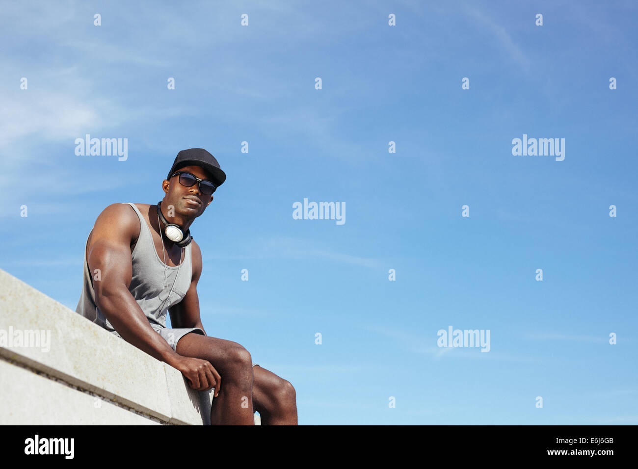Elegante giovane africano uomo seduto contro il cielo blu. Uomo che indossa gli occhiali da sole e cappello con un paio di cuffie guardando la fotocamera. Foto Stock