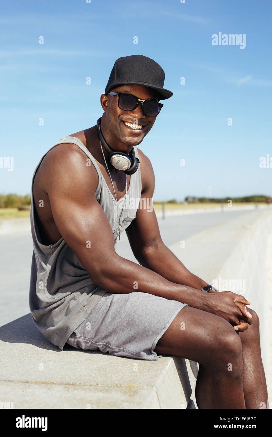 Ritratto di felice del giovane africano seduto su una passeggiata. Elegante modello maschile di indossare occhiali da sole e cappello. Foto Stock