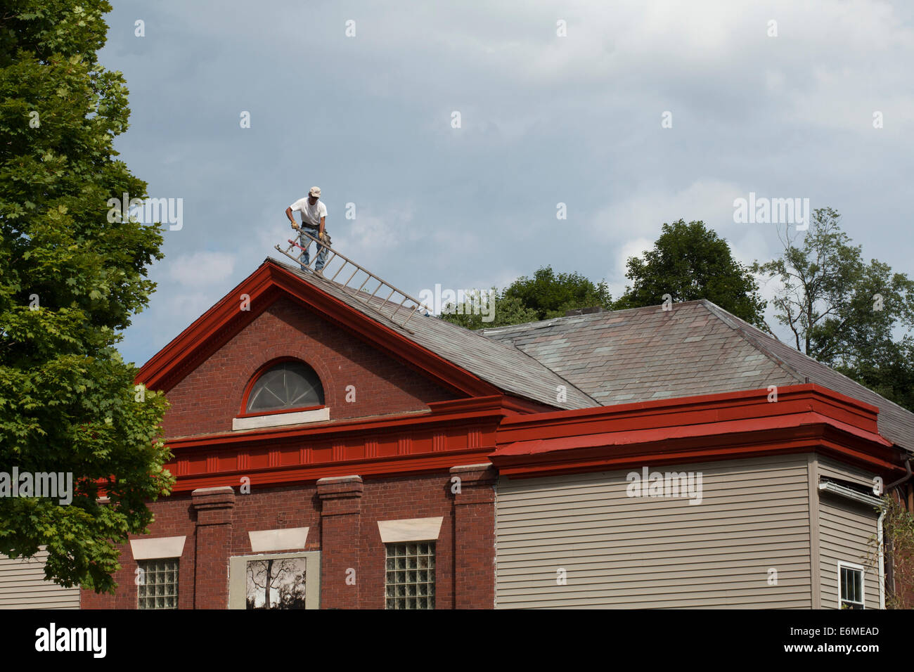 Un copritetti muove una scaletta durante la riparazione di un vecchio tetto in ardesia su un ex schoolhouse. Foto Stock
