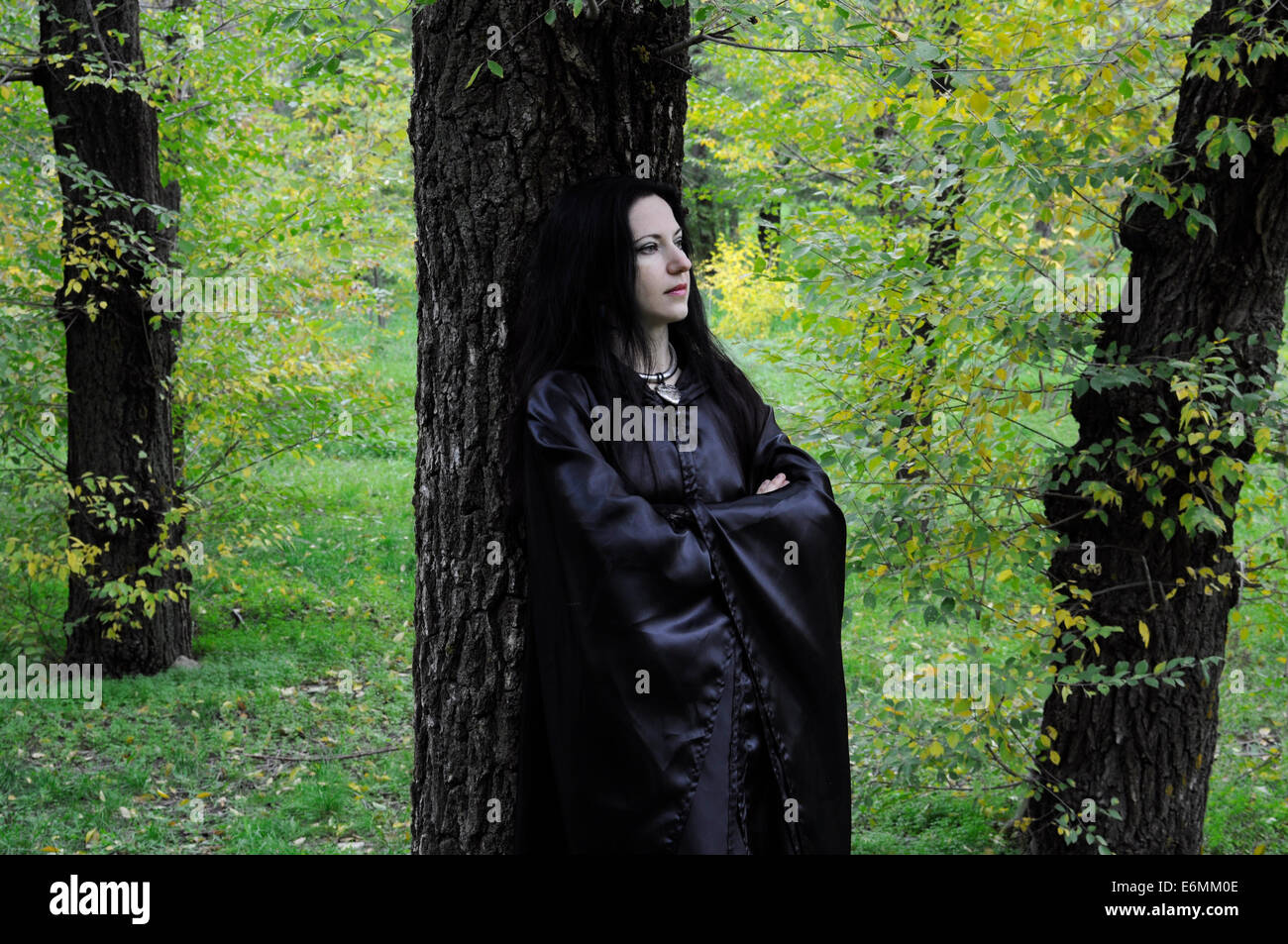 La donna in nero in piedi contro un oscuro tronco di albero nel verde della foresta di autunno Foto Stock