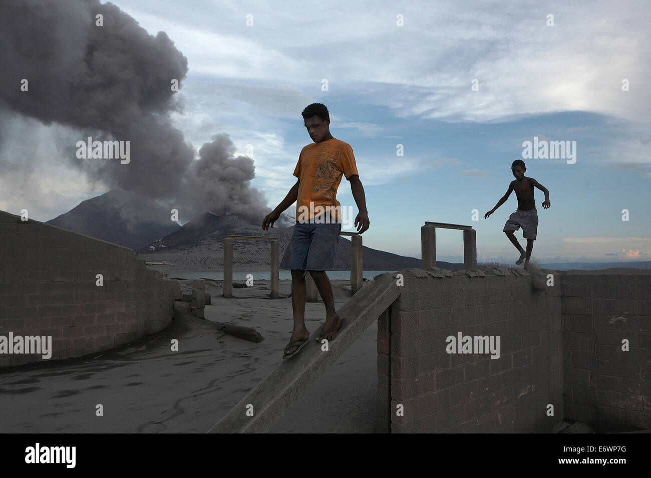 Bambini che giocano tra le rovine di una vecchia fabbrica di cemento con il vulcano in eruzione in background. Vulcano Tavurvur, Rabaul, est n Foto Stock