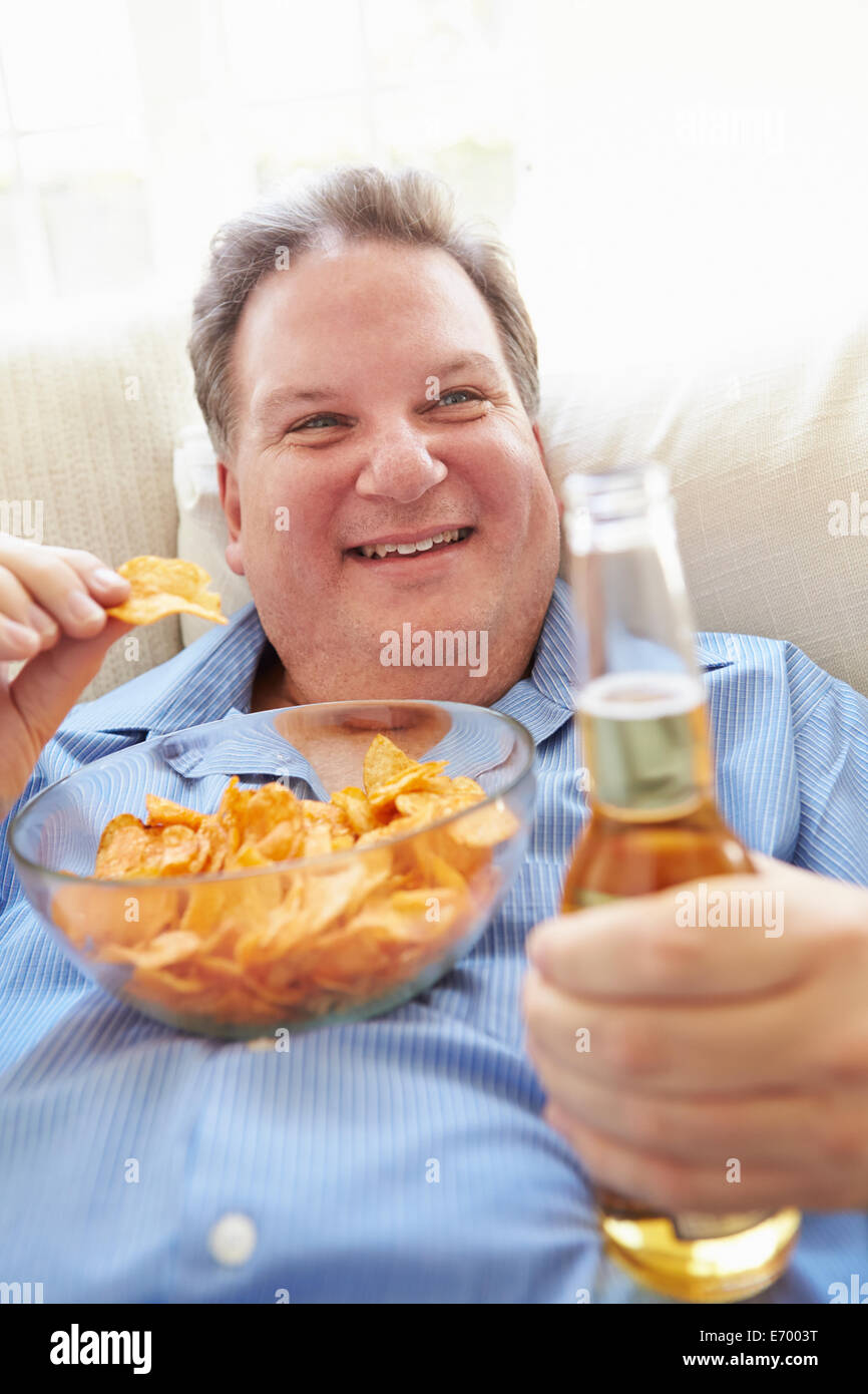 Uomo sovrappeso a casa a mangiare patatine e bere birra Foto Stock