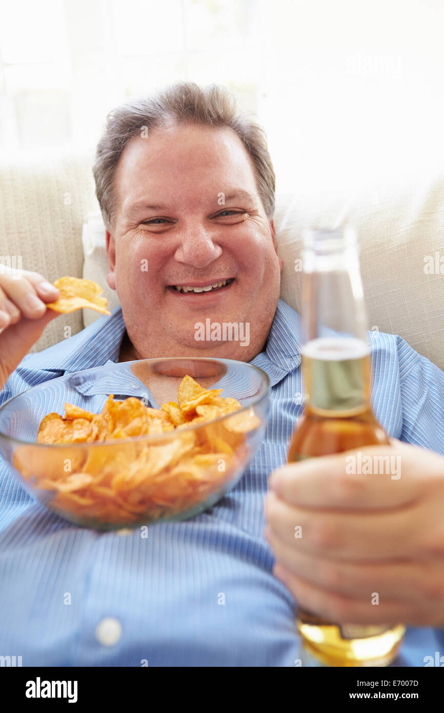 Uomo sovrappeso a casa a mangiare patatine e bere birra Foto Stock