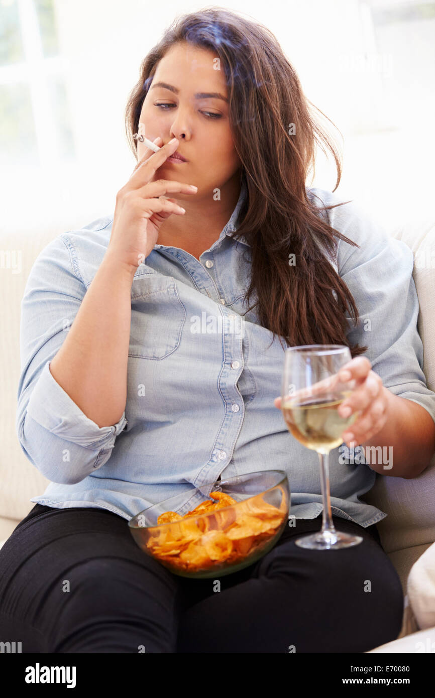 Donna sovrappeso mangiare patatine, bere vino e fumare Foto Stock