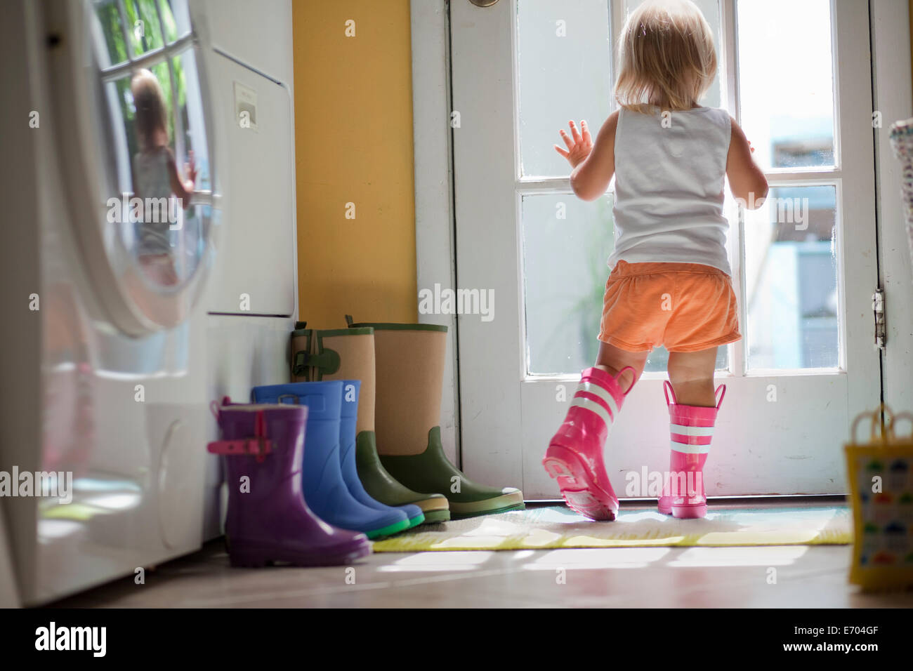 Toddler femmina indossare stivali di gomma guardando fuori della porta finestra Foto Stock