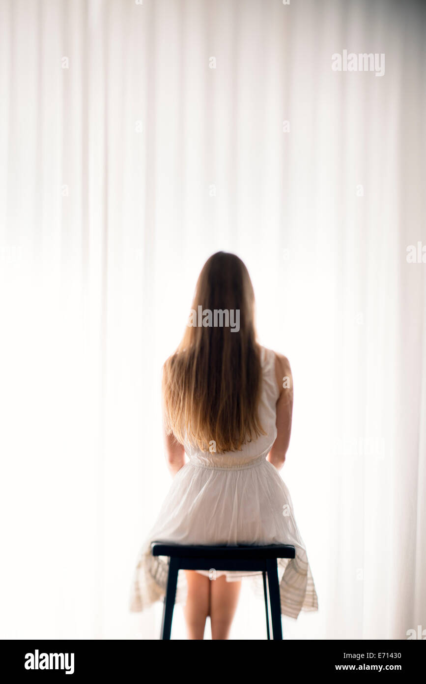 Giovane donna seduta su uno sgabello davanti a una tenda bianca, vista posteriore Foto Stock