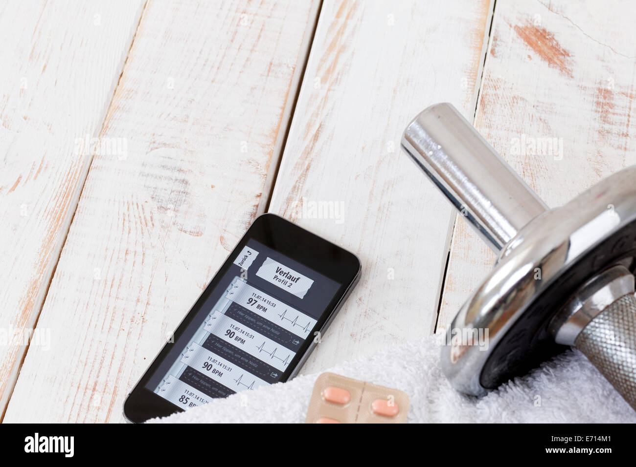 Smartphone mostra monitor frequenza cardiaca, asciugamano, compresse e dumbell su legno Foto Stock