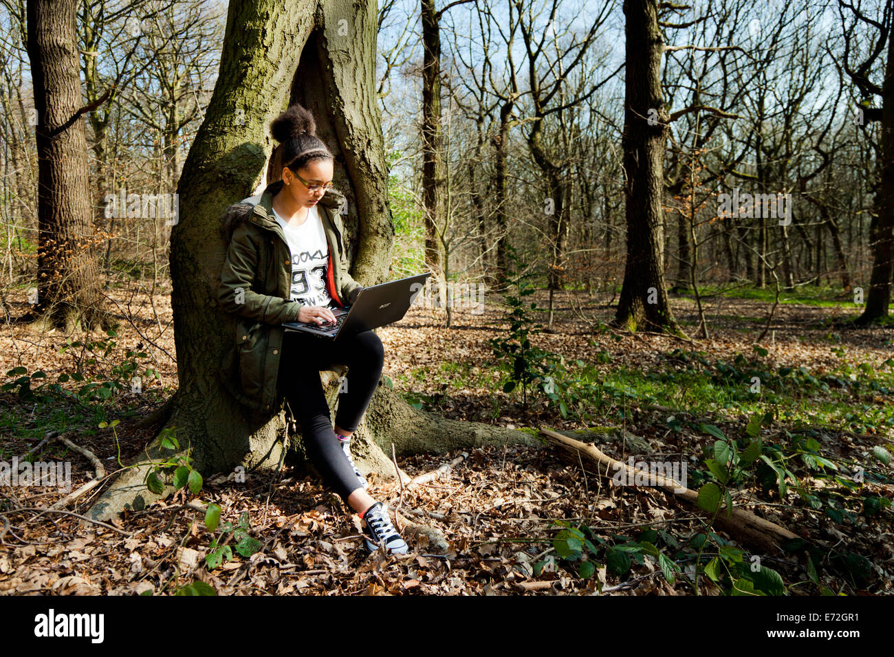 Giovani razza mista ragazza si appoggia contro un albero in una foresta e accede al suo computer portatile. Foto Stock
