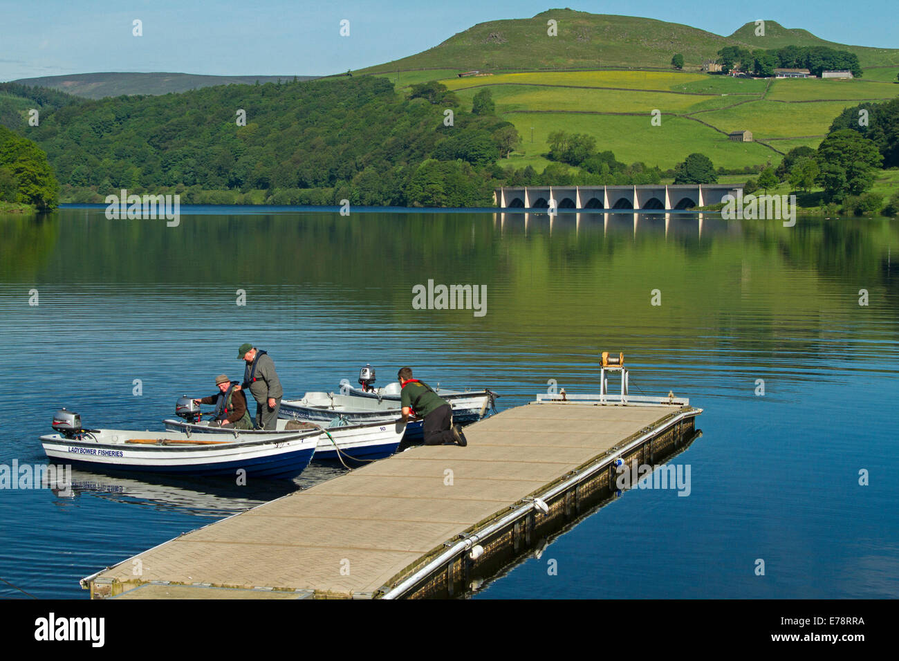 I pescatori in colata in barca dal molo al Lago Ladybower con calma acqua blu ai piedi della collina con boschi e terreni coltivati in Peak National Park in Inghilterra Foto Stock