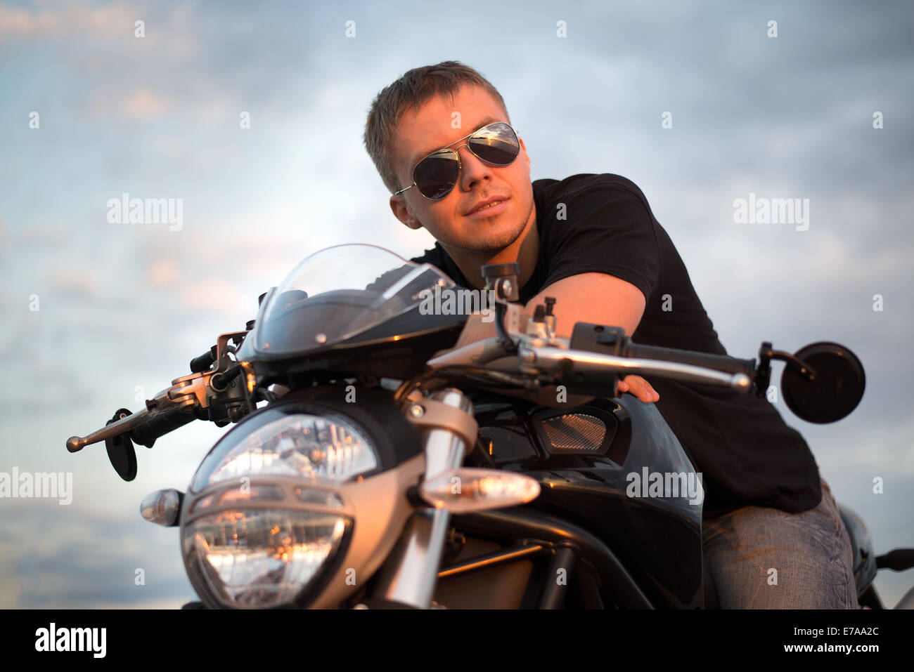 Ritratto romantico bello biker uomo in occhiali da sole si siede su una bicicletta su un tramonto vicino al lago e la città Foto Stock