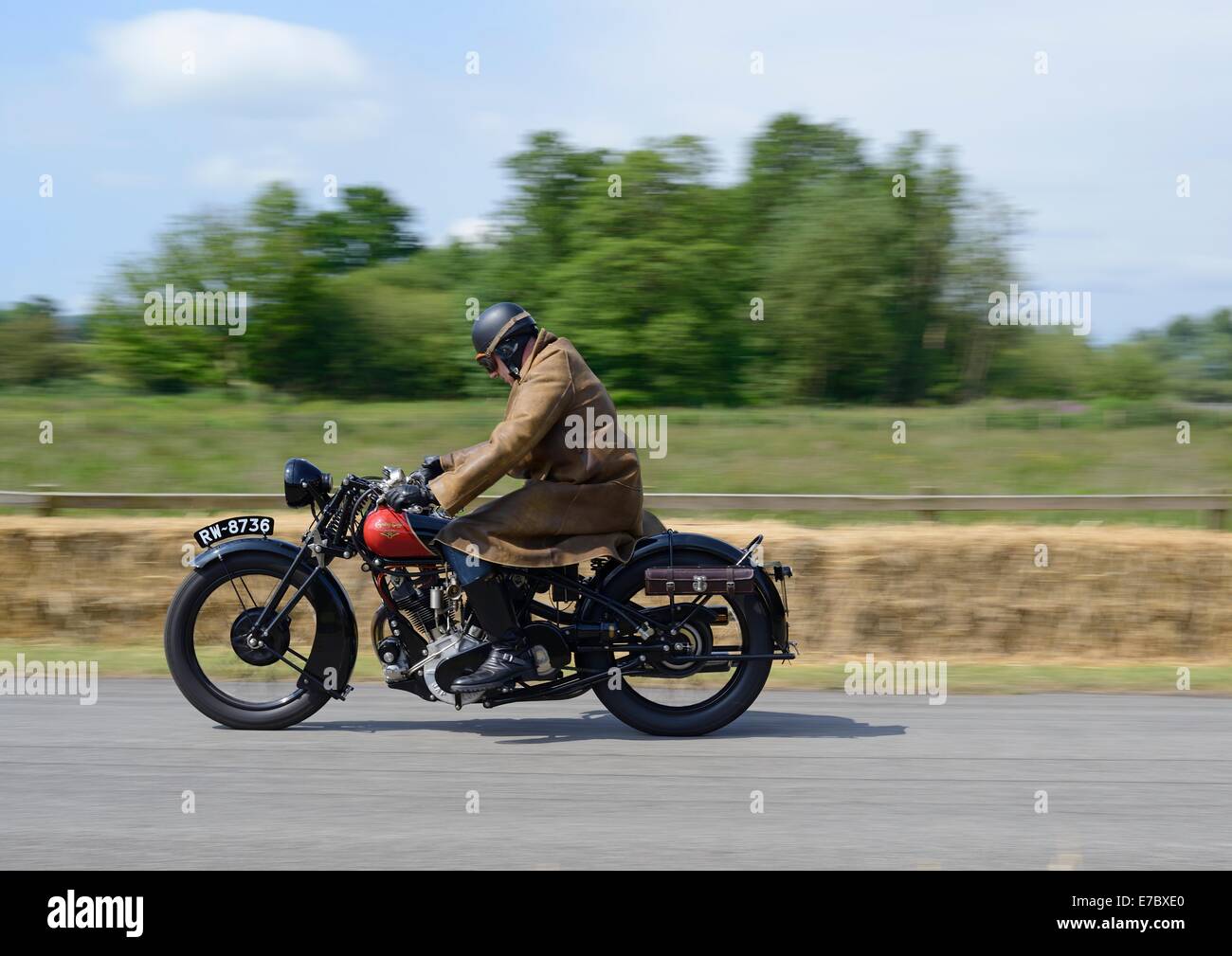 Uomo in cappotto lungo, casco e occhiali in sella a una motocicletta vintage, balle di fieno dietro Foto Stock