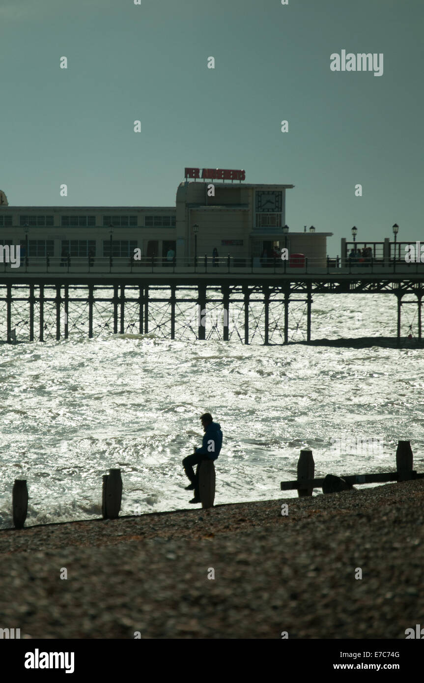 Un uomo si siede sul mare difese a Worthing con il molo in background Foto Stock