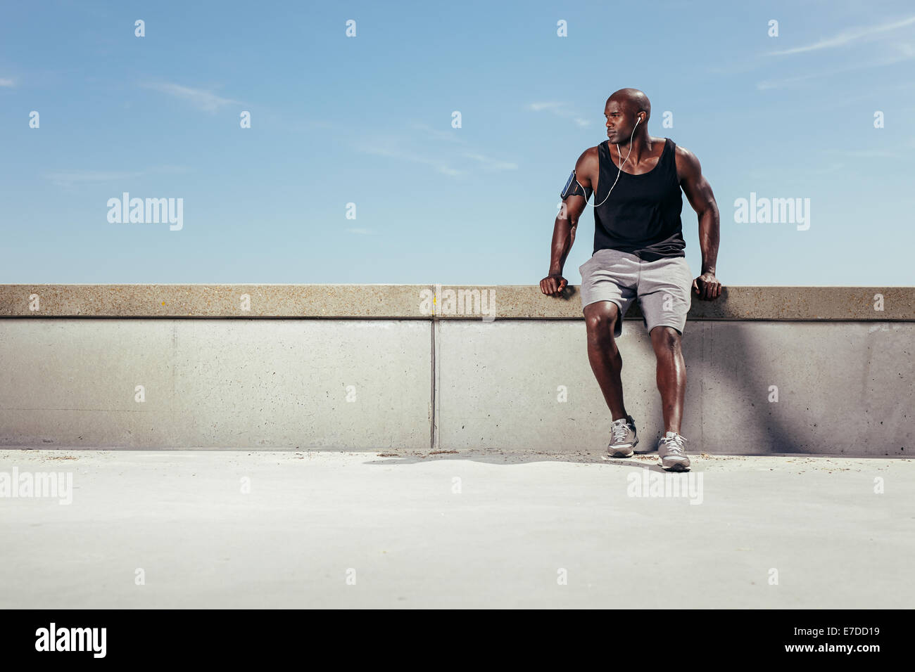 Muscoloso giovane rilassante sul terrapieno che guarda lontano copyspace. Maschio africano atleta che indossa gli auricolari all'esterno. Maschio di runner. Foto Stock