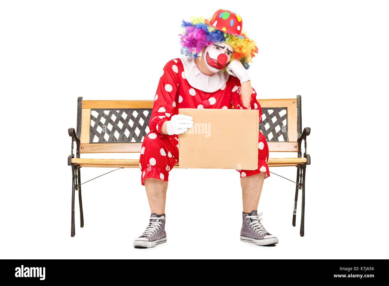 Triste clown maschile seduto su una panca in legno e in possesso di un cartone vuoto segno isolato su sfondo bianco Foto Stock