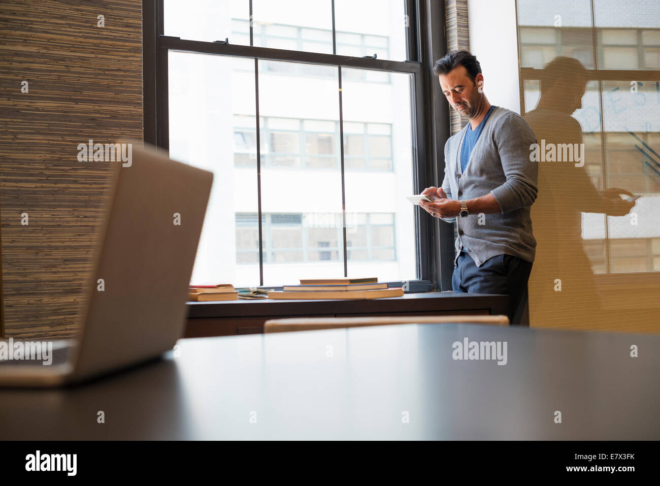 Vita in ufficio. Un uomo in piedi da una finestra in un ufficio controllo il suo smart phone. Foto Stock