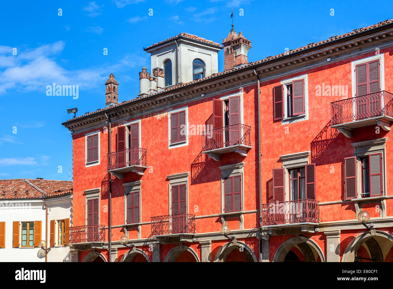 La facciata della red edificio a due piani con balconi e persiane di legno sotto il cielo blu di Alba, Italia. Foto Stock