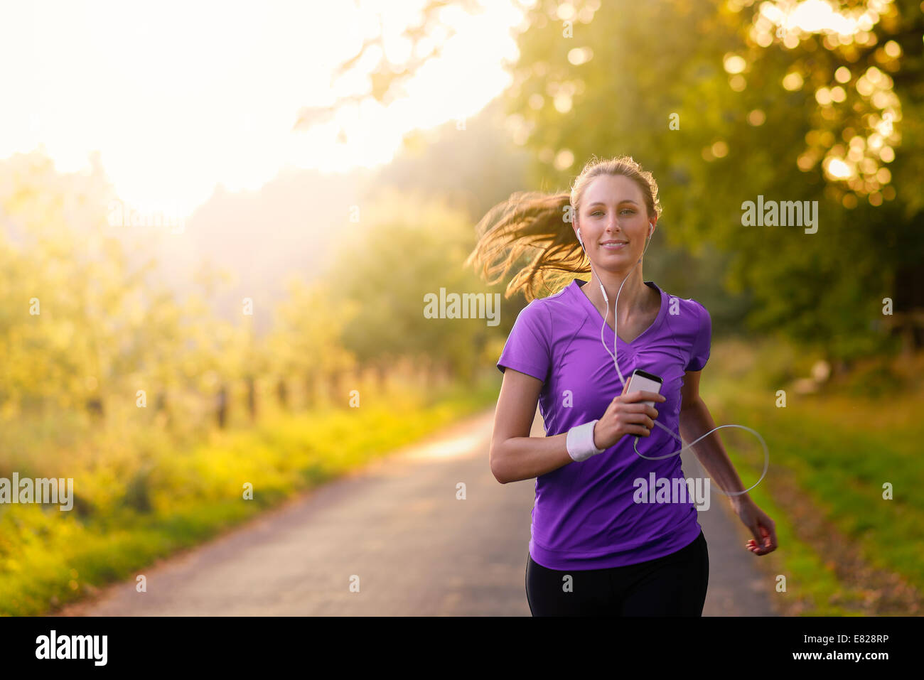 Donna ascoltando la sua musica su i tappi per le orecchie e un lettore MP3 durante il jogging lungo una strada di campagna in uno stile di vita sano, esercizio e Foto Stock