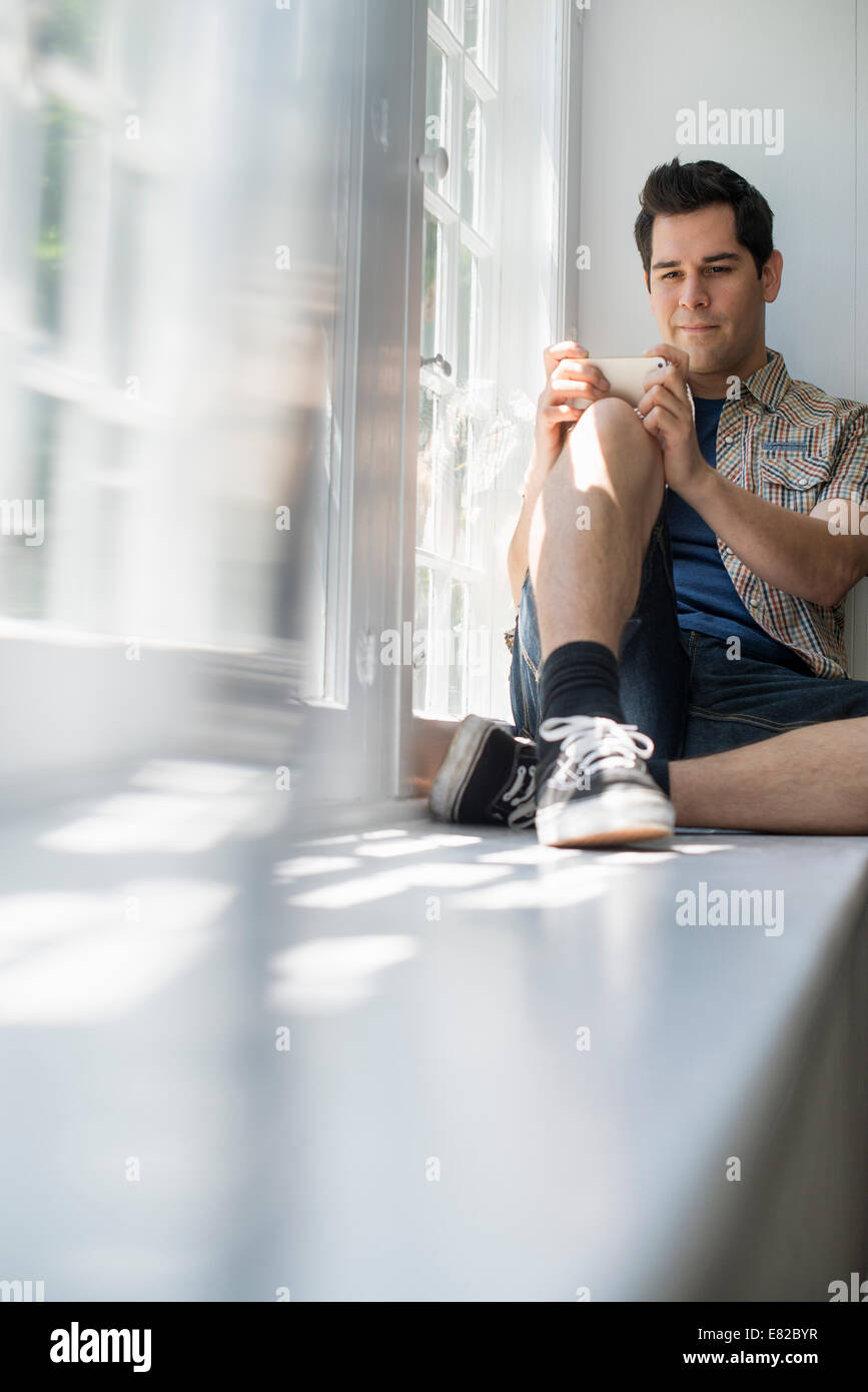 Uomo seduto da una finestra, tenendo in mano un telefono cellulare. Foto Stock