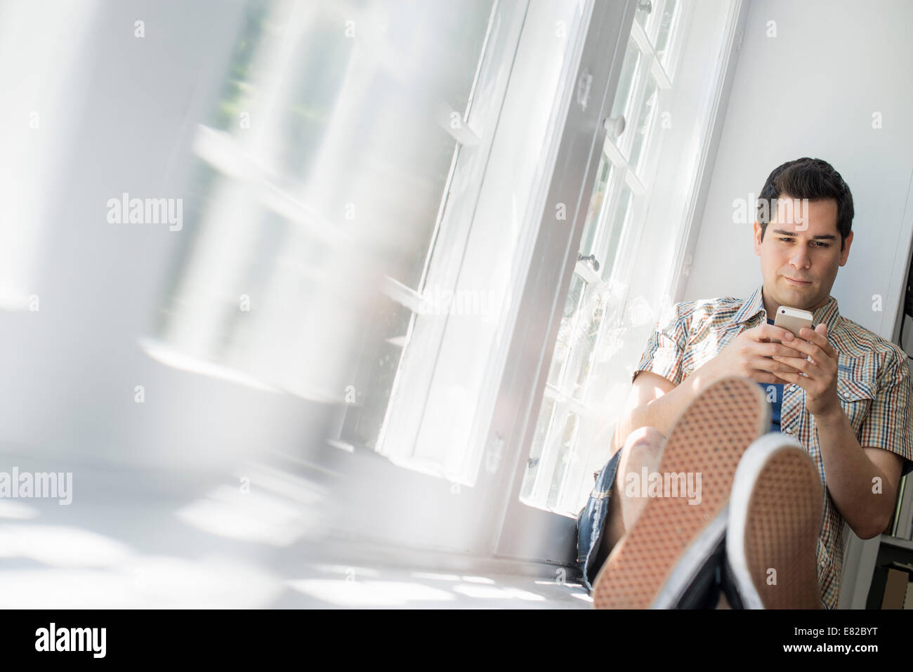 Uomo seduto da una finestra, tenendo in mano un telefono cellulare. Foto Stock