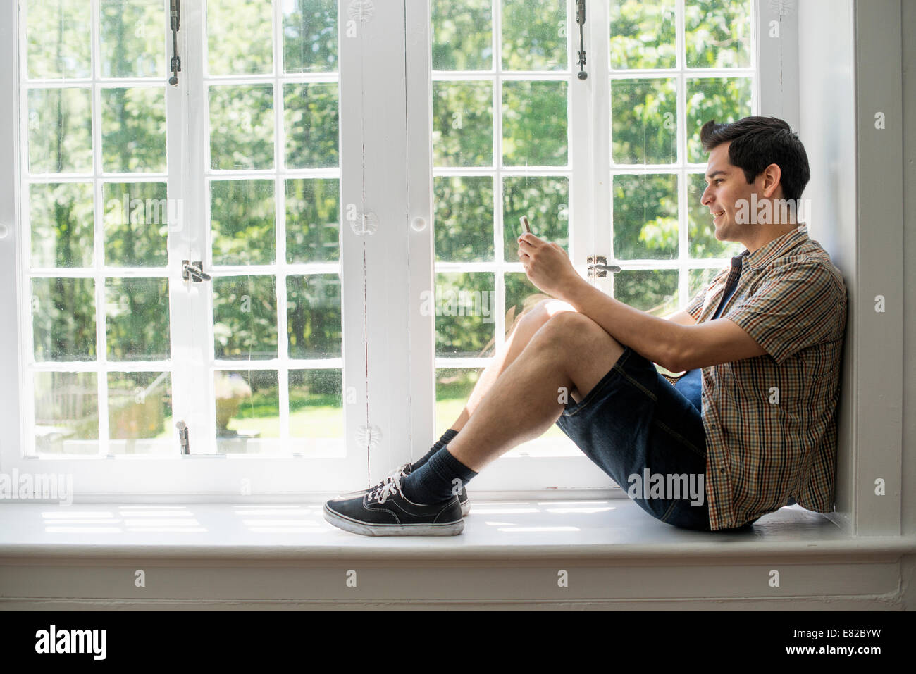 Uomo seduto da una finestra, tenendo in mano un telefono cellulare. Foto Stock