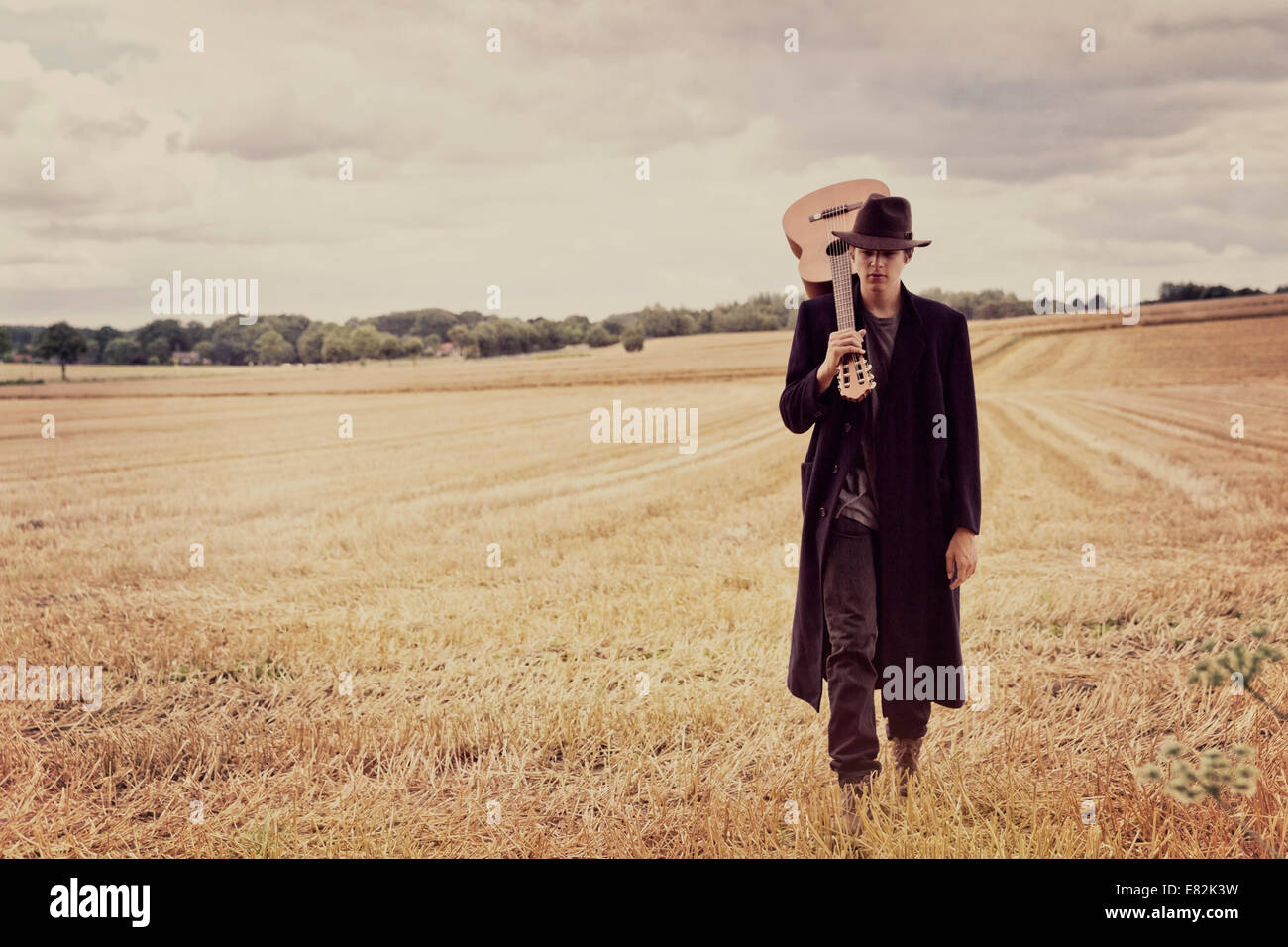 Ragazzo adolescente con il cappello e il cappotto lungo camminare su un grainfield con la chitarra Foto Stock