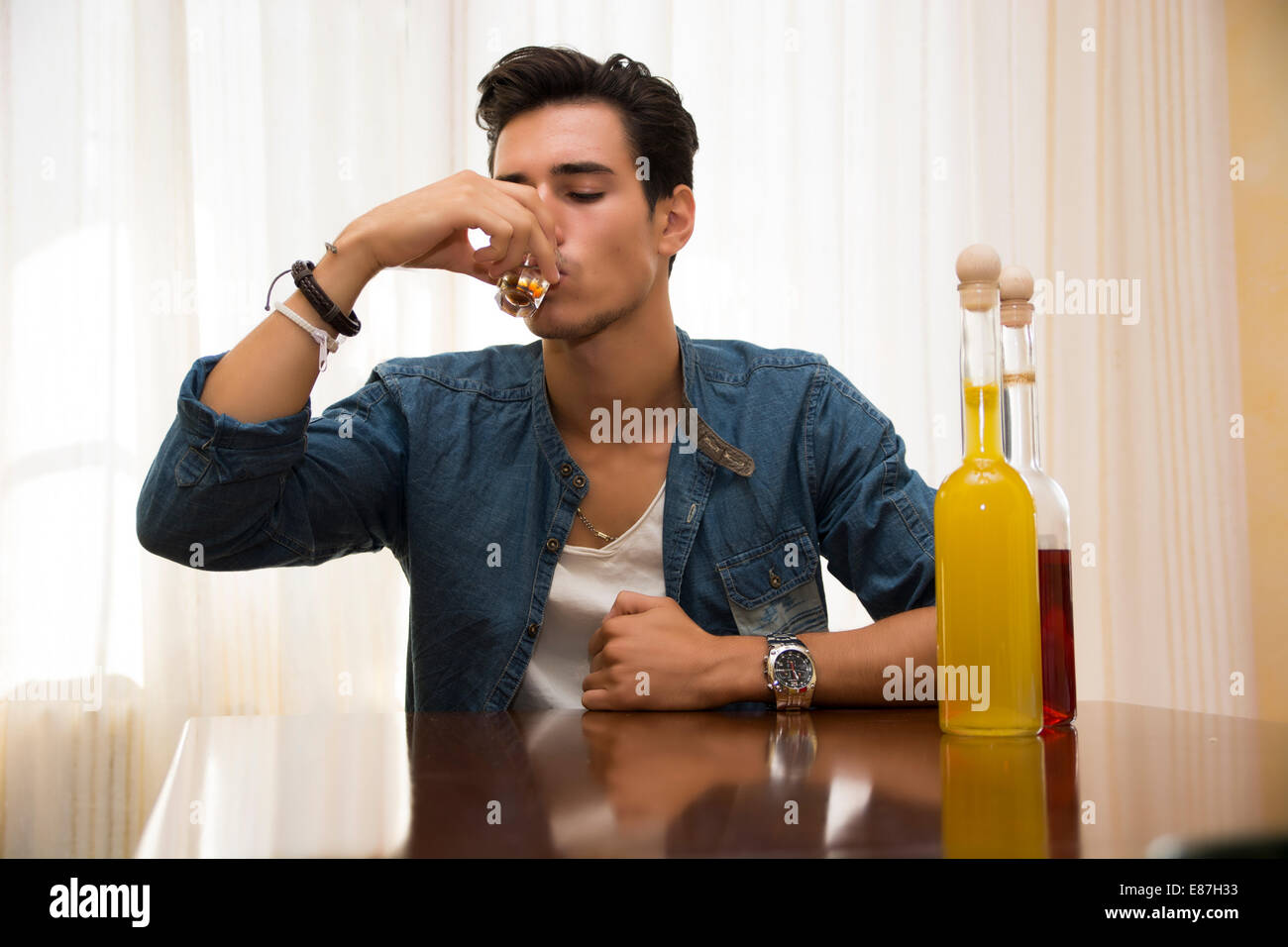 Giovane uomo seduto a bere da solo ad un tavolo con due bottiglie di liquori accanto lui sorseggiando dal colpo di vetro di affogare il suo dolore Foto Stock