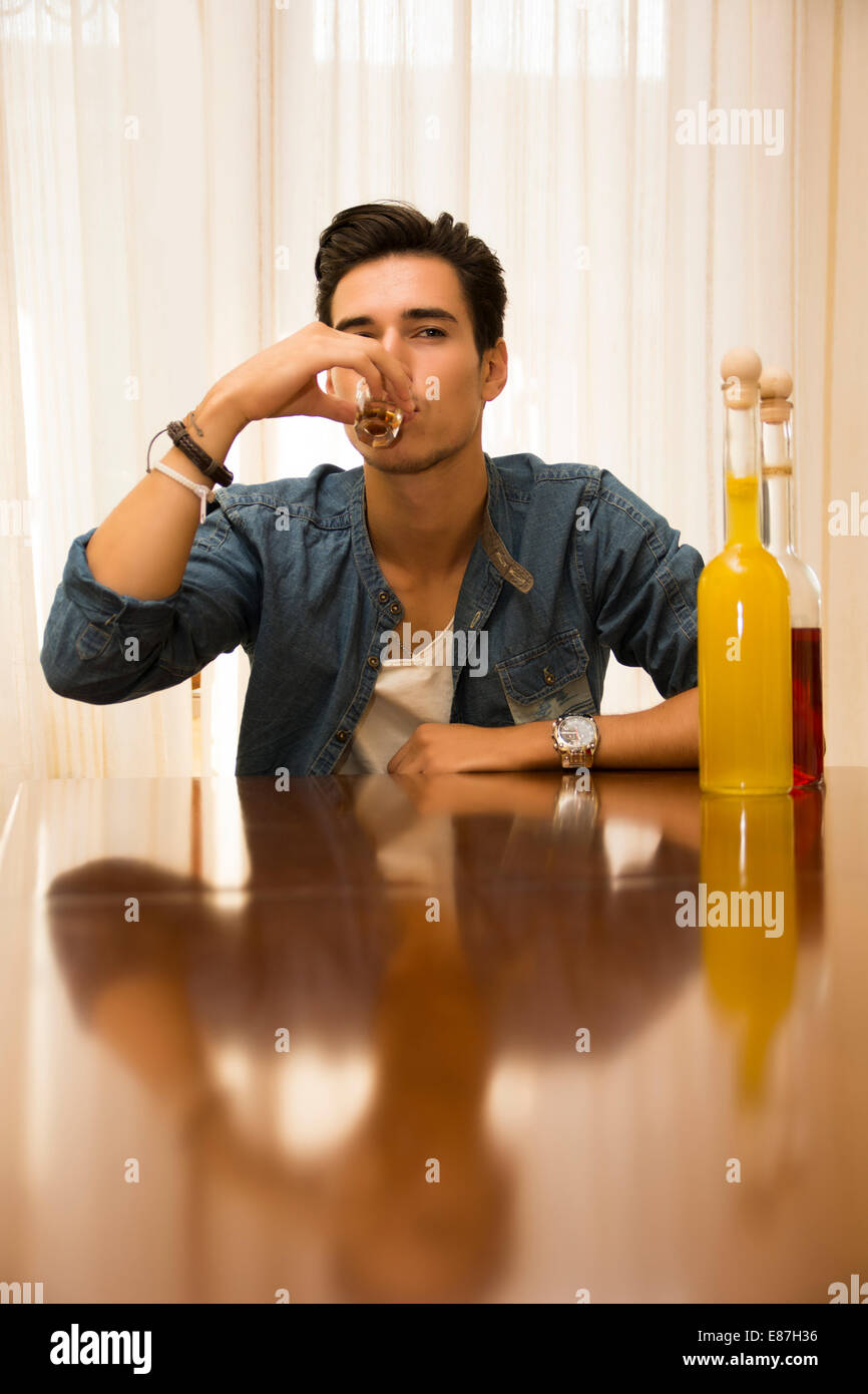 Giovane uomo seduto a bere da solo ad un tavolo con due bottiglie di liquori accanto lui sorseggiando dal colpo di vetro di affogare il suo Foto Stock