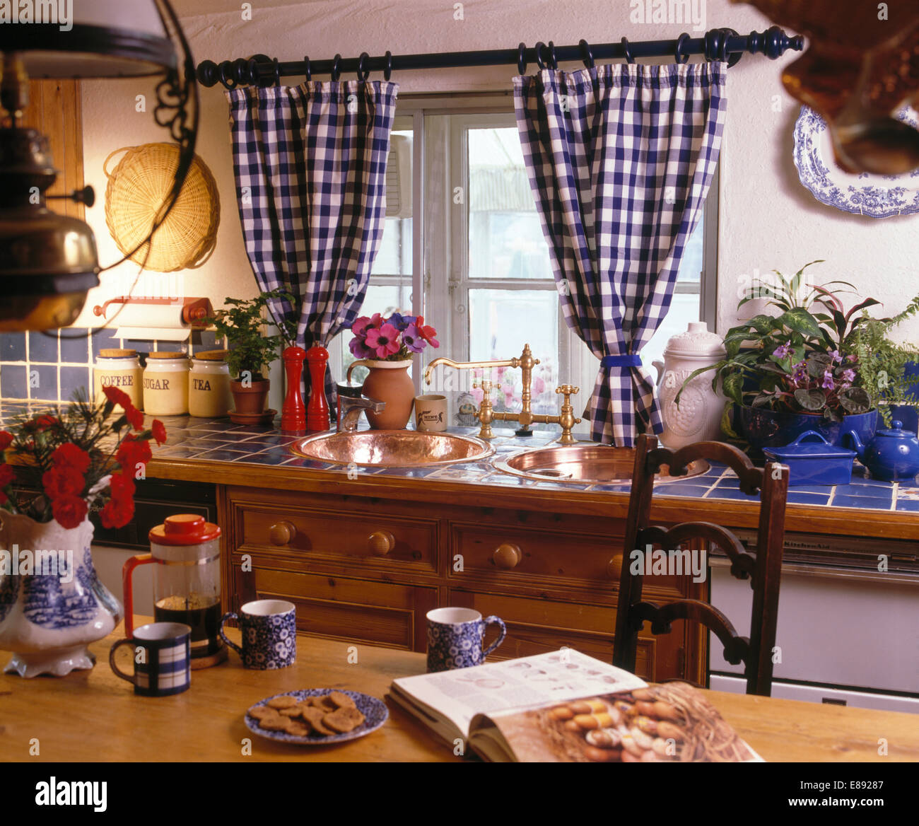 Blue+bianco tende selezionata sulla finestra in cottage con cucina cafetiere rosso e blu tazze sul tavolo Foto Stock