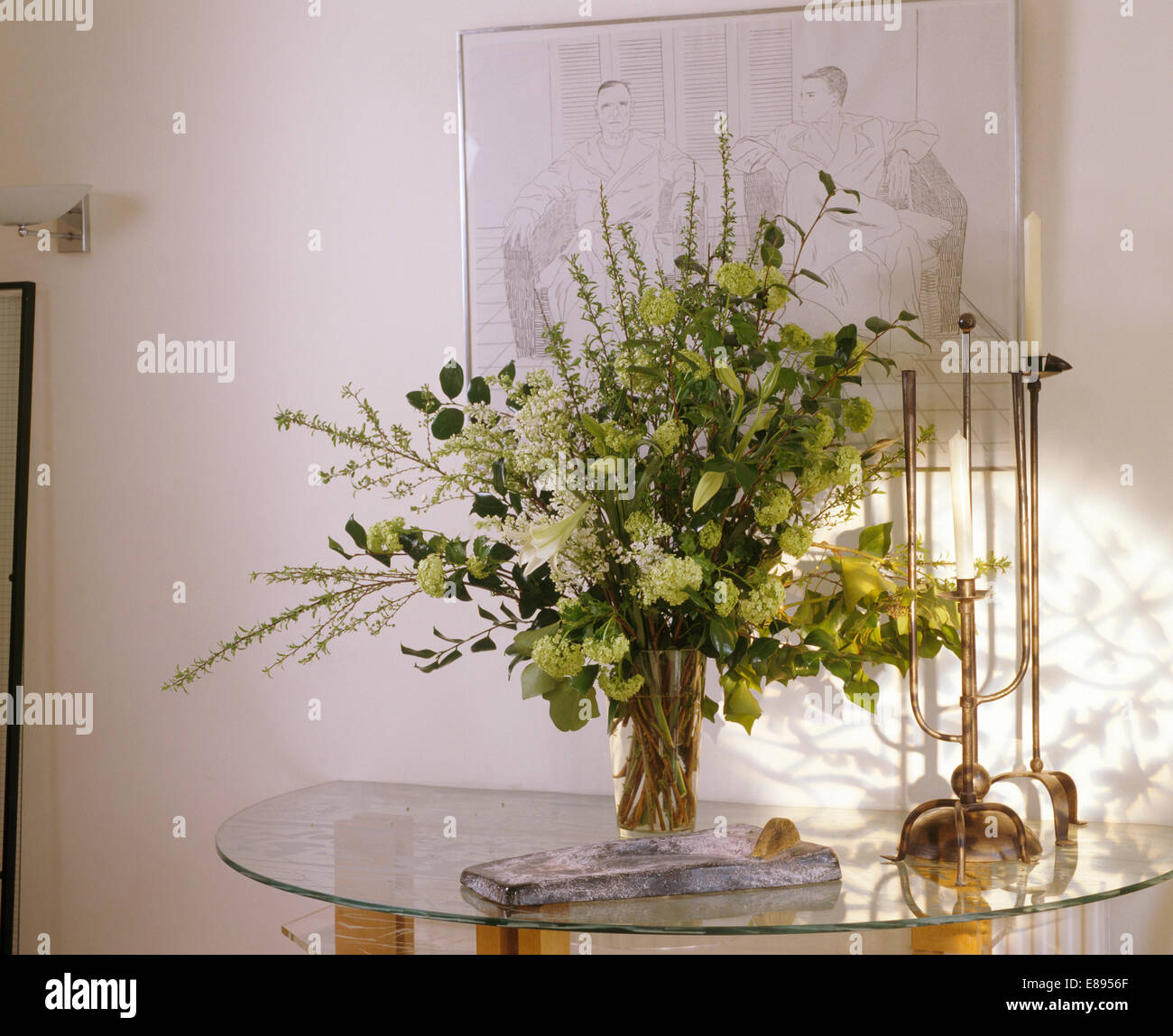 Close-up di fiori bianchi e foglie verdi in vaso di vetro su vetro tavolo console con alte candeliere Foto Stock