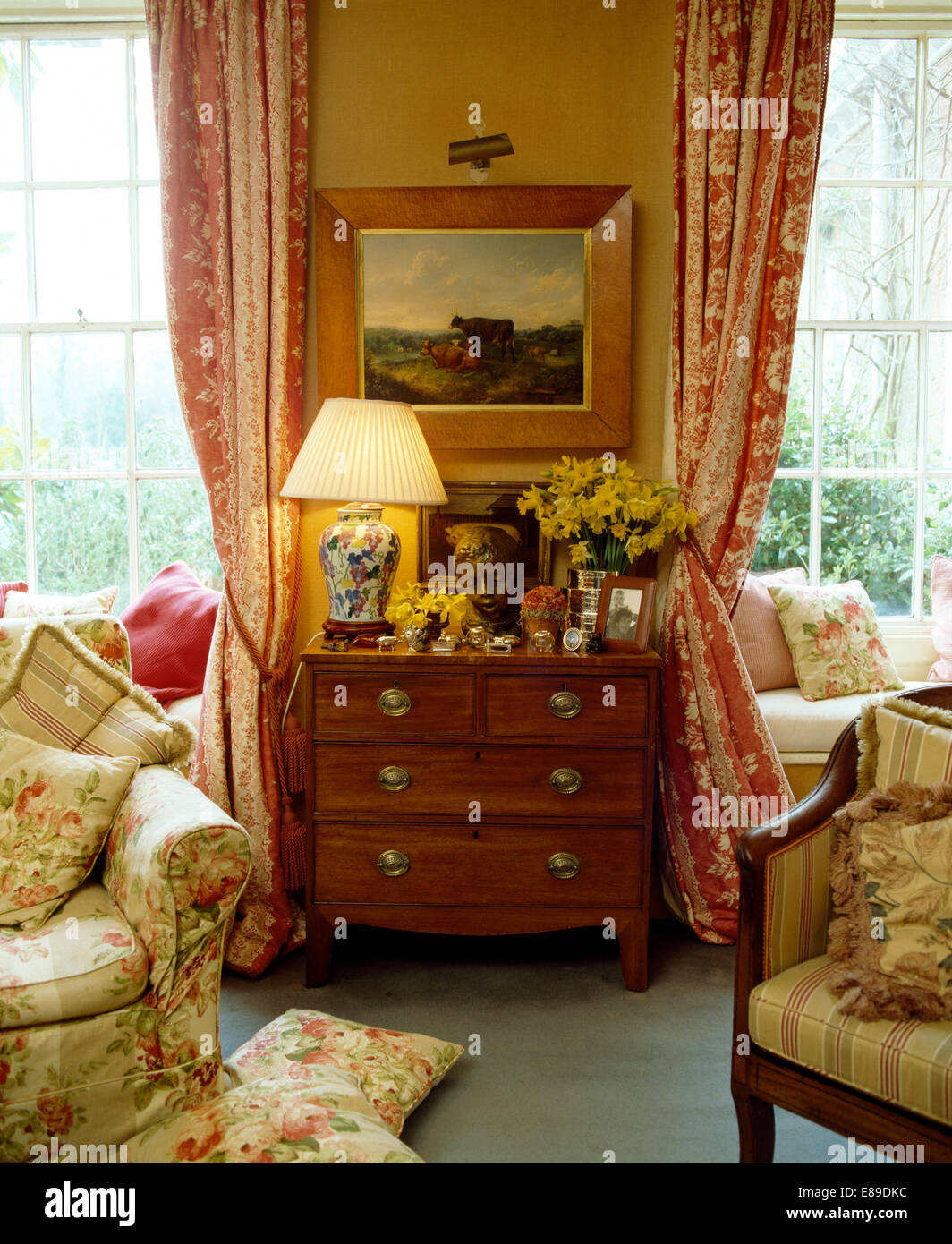 La pittura di paesaggio al di sopra di torace-di-cassetti con lampada accesa nel paese soggiorno con poltrona floreali e tendaggi rossi Foto Stock