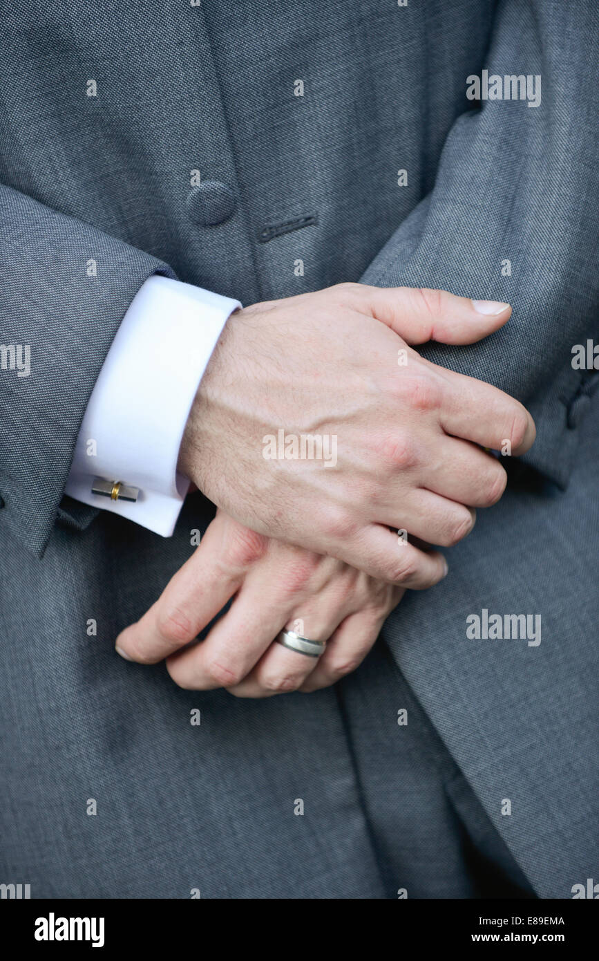 Le due mani incrociate di un uomo caucasico indossando un tradizionale grigio tight, camicia bianca, polsini e un anello di nozze Foto Stock