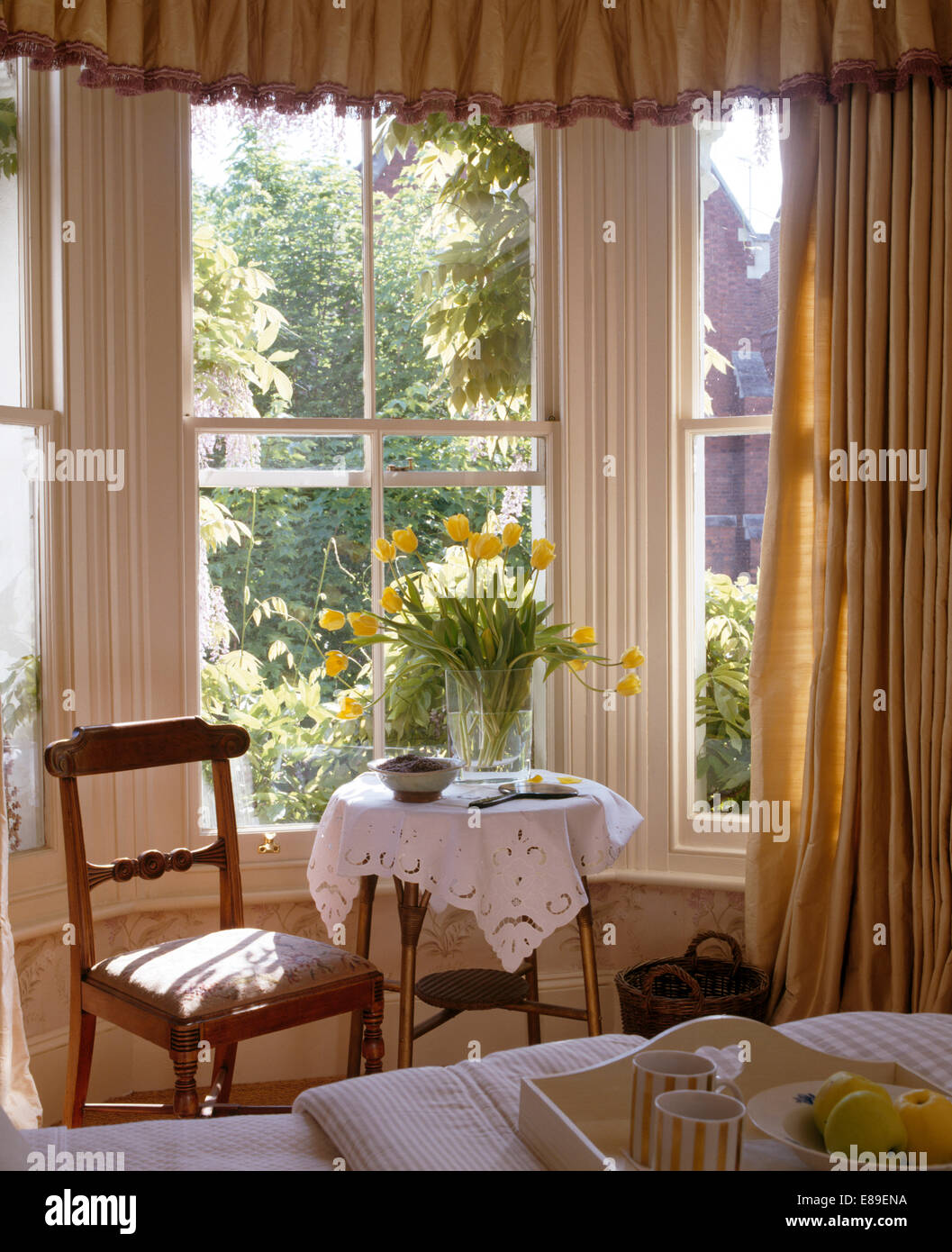 Sedia antica e un piccolo tavolo con broderie anglaise tessuto nella parte anteriore della finestra di baia con pesanti tende di seta Foto Stock