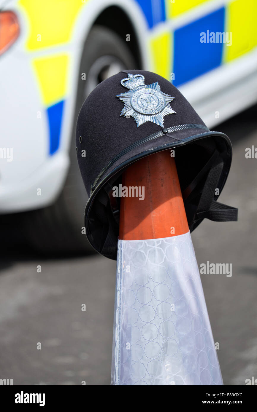 Un britannico poliziotti casco tradizionale appollaiato su un traffico cono contro lo sfondo di una de-focalizzata auto della polizia. Foto Stock