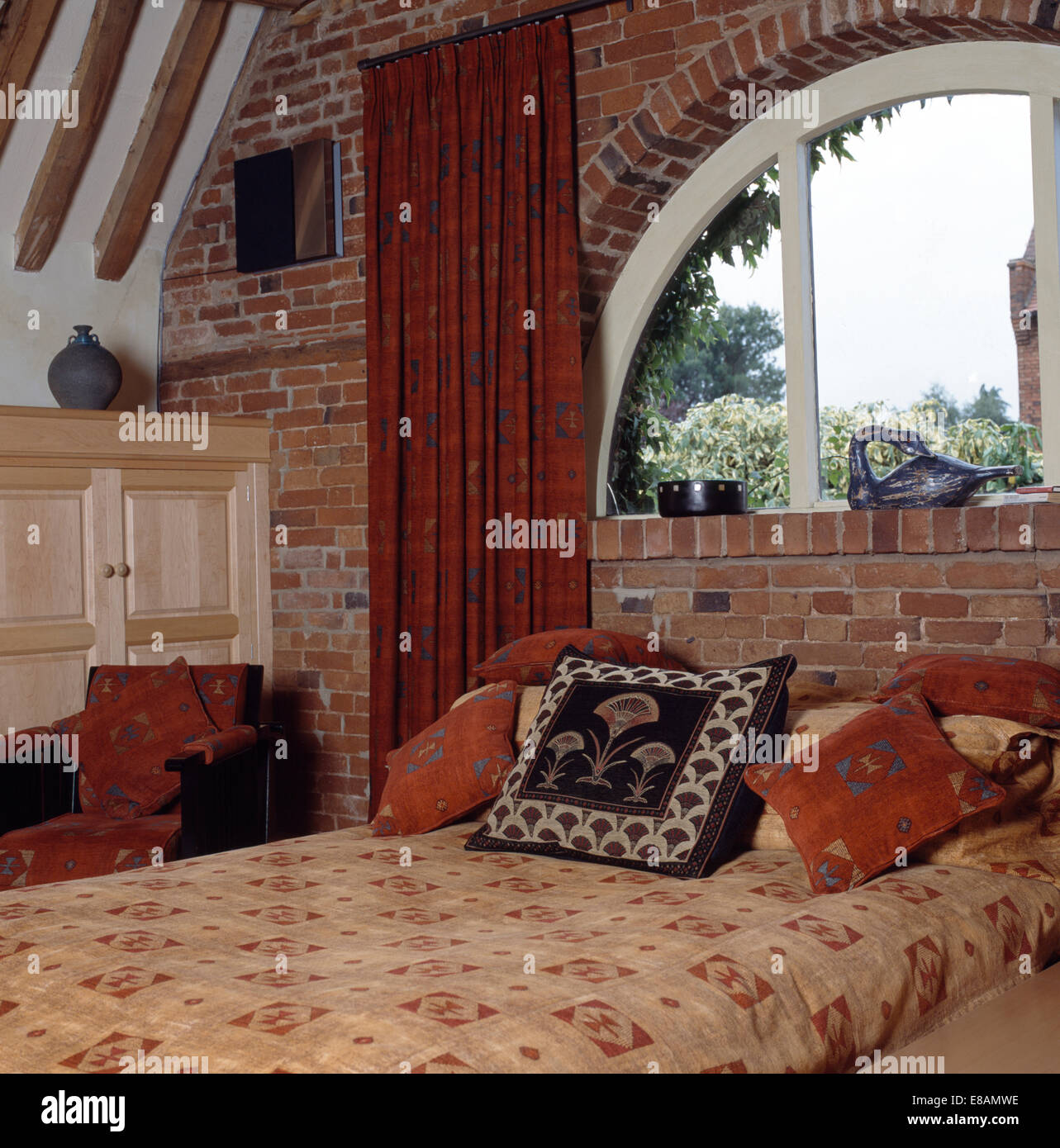 Modellato cuscini e piumoni sul letto sotto la finestra ad arco con tende rosse nel fienile nella camera da letto di conversione Foto Stock