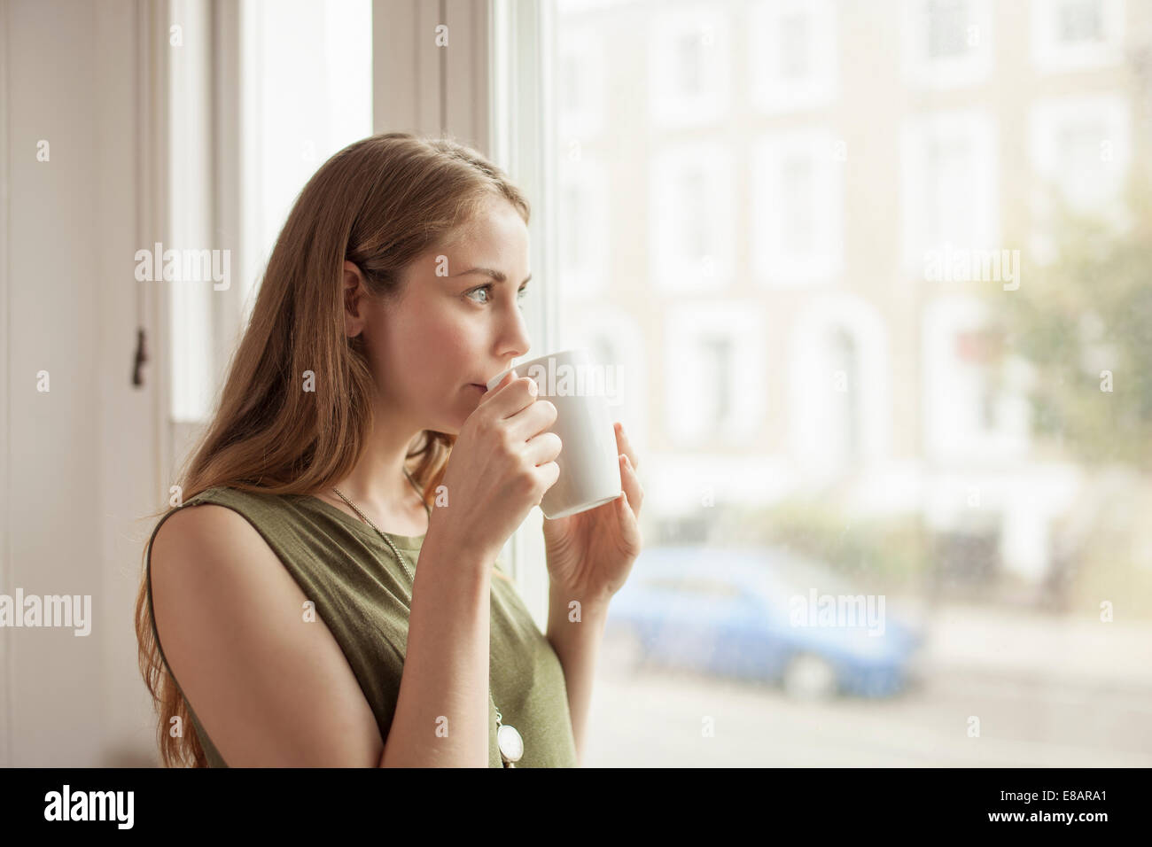 Giovane donna di bere il caffè e guardare attraverso la stanza di seduta finestra Foto Stock