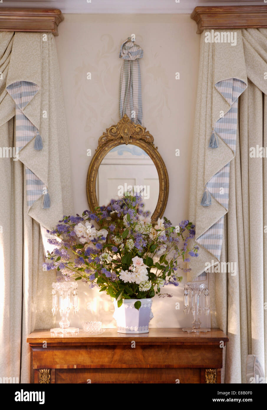 Antico specchio dorato al di sopra di un vaso di fiori freschi sul tavolo piccolo tra la crema e striato swagged tende Foto Stock