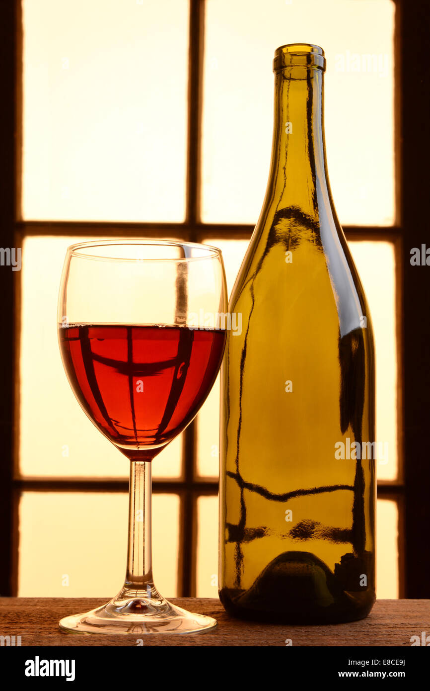 Un vino ancora in vita con toni caldi. Un vuoto di bottiglie di vino e un bicchiere di vino rosso davanti a una finestra con una calda luce del sole. Foto Stock