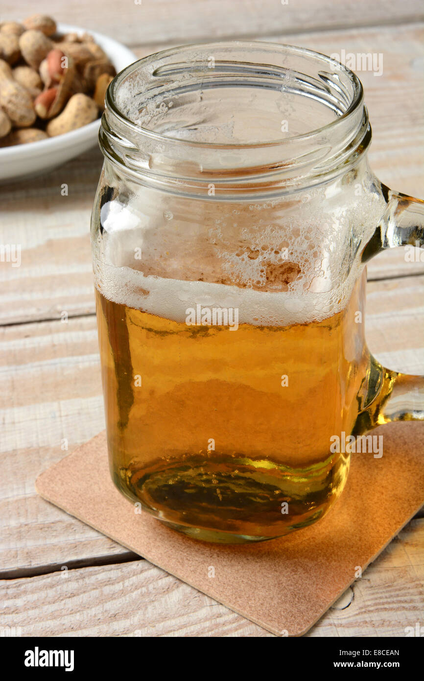 Primo piano di un vasetto mug con la birra. Formato verticale su una tavola in legno rustico e arachidi in background. Foto Stock