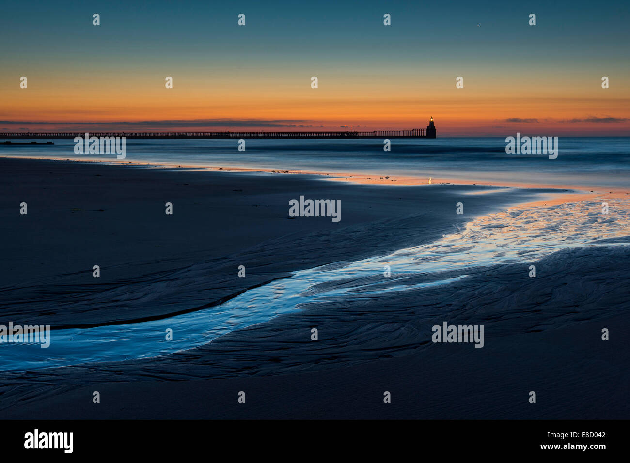 La mattina presto a Blyth. Linea creata dalla riflessione sull'acqua porta verso il mare, il faro e il molo. Foto Stock