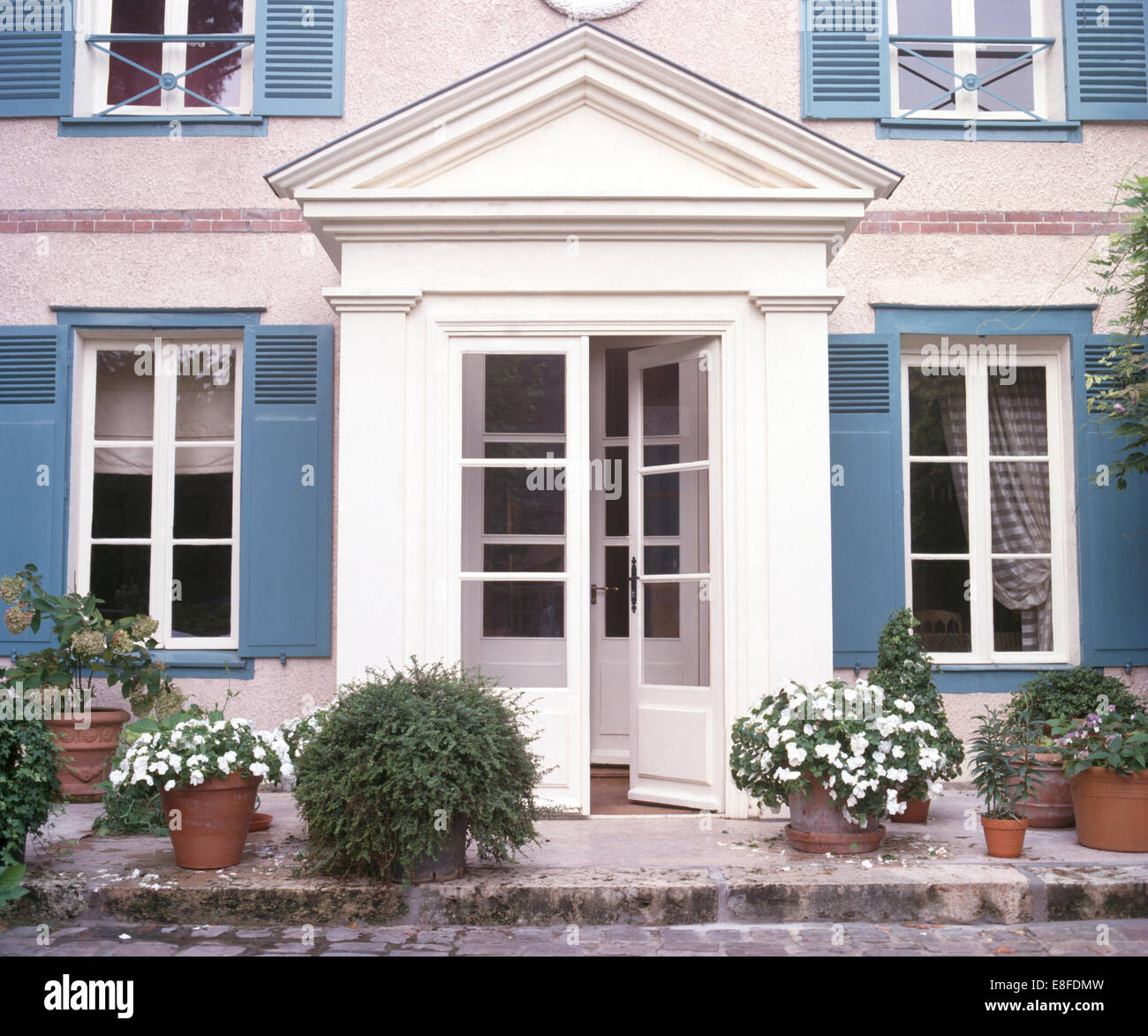 Impatiens bianco in vasi su entrambi i lati del mezzo vetri delle porte anteriori della casa francese di campagna con persiane blu Foto Stock