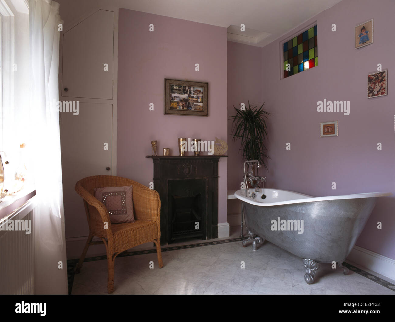Verniciato argento roll top bagno in viola pallido bagno con sedia di vimini accanto al caminetto Foto Stock