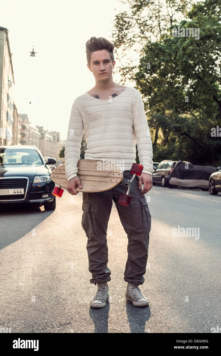A piena lunghezza verticale maschio fiducioso studente di scuola superiore con lo skateboard in piedi su una strada di città Foto Stock