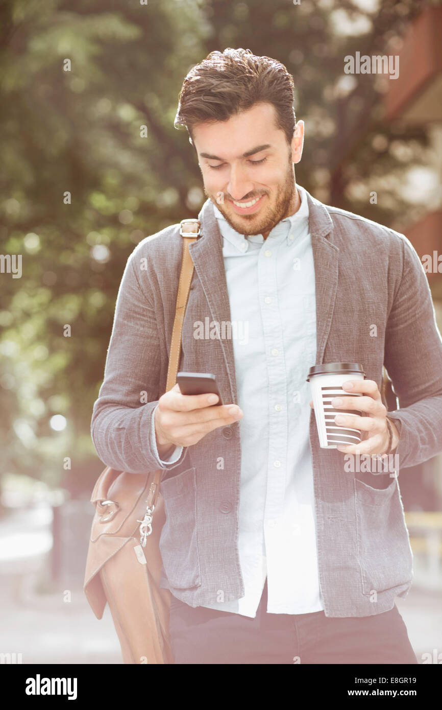 Imprenditore sorridente un caffè durante la lettura di un messaggio di testo sul telefono intelligente all'aperto Foto Stock