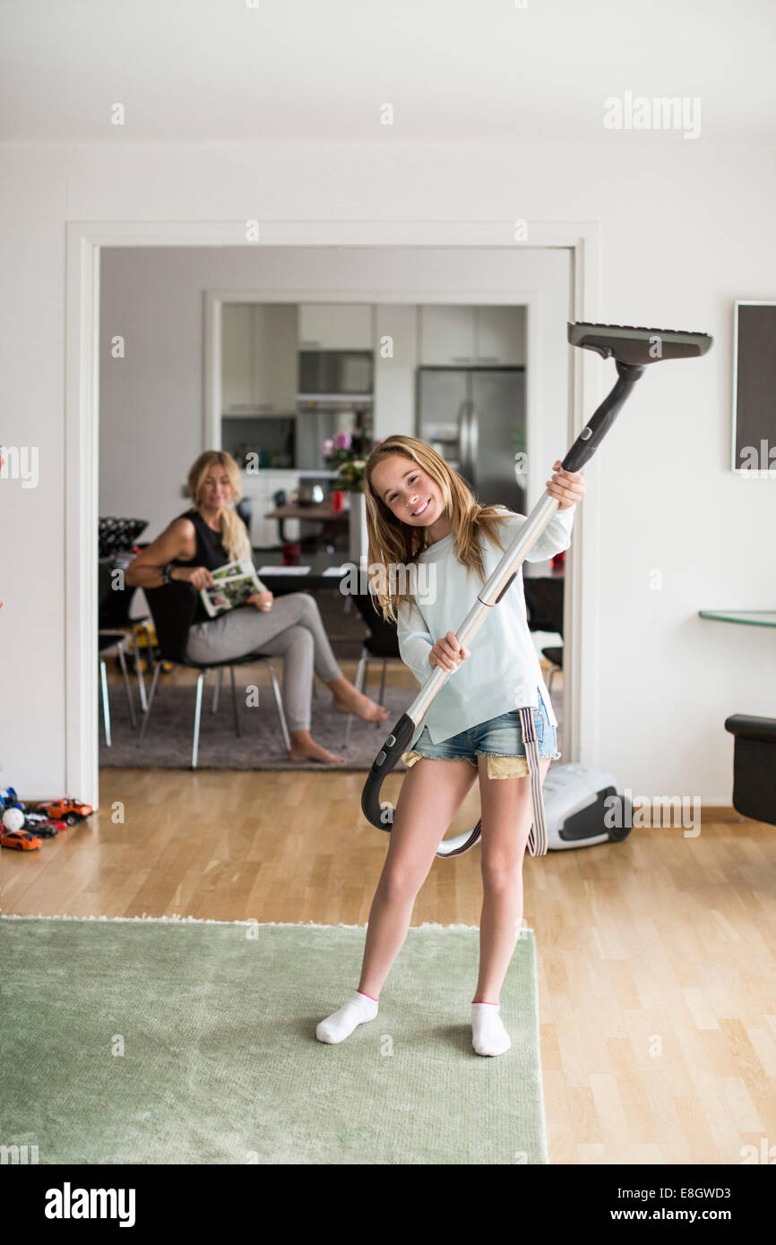 A piena lunghezza Ritratto di giocoso ragazza con aspirapolvere a casa con la madre seduta in background Foto Stock