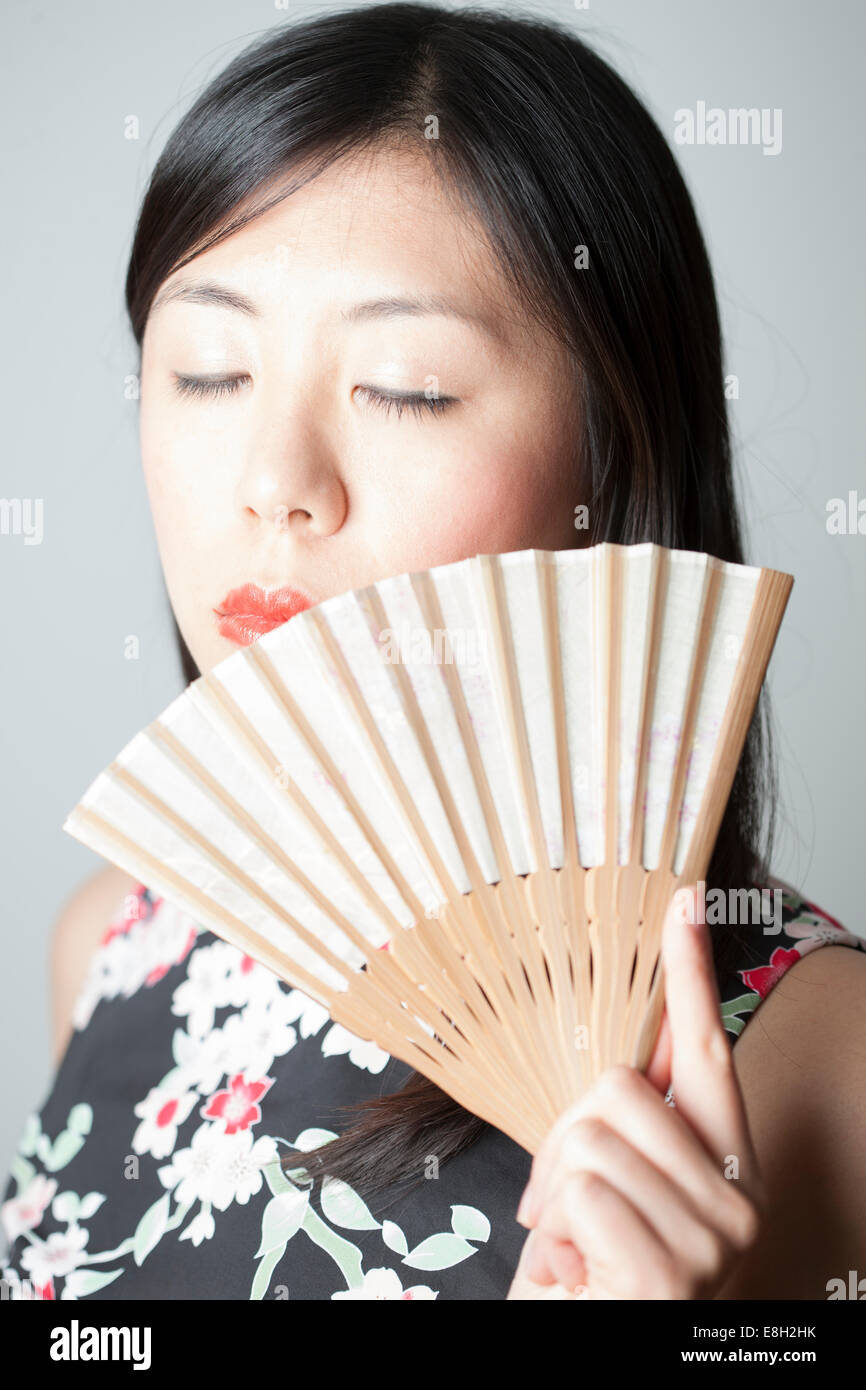 Ritratto di donna asiatica con gli occhi chiusi mantenendo la ventola davanti a uno sfondo grigio Foto Stock