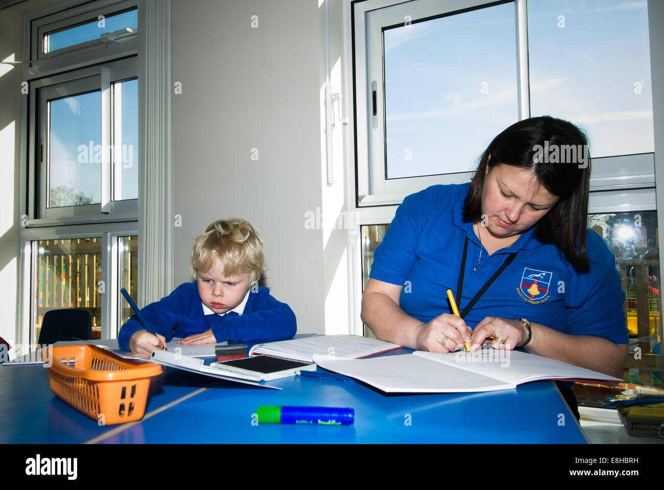 Bambino in pre-scuola di classe con Learning Support Assistant (LSA), Oxfordshire, Regno Unito iscritto in un libro di scuola Foto Stock