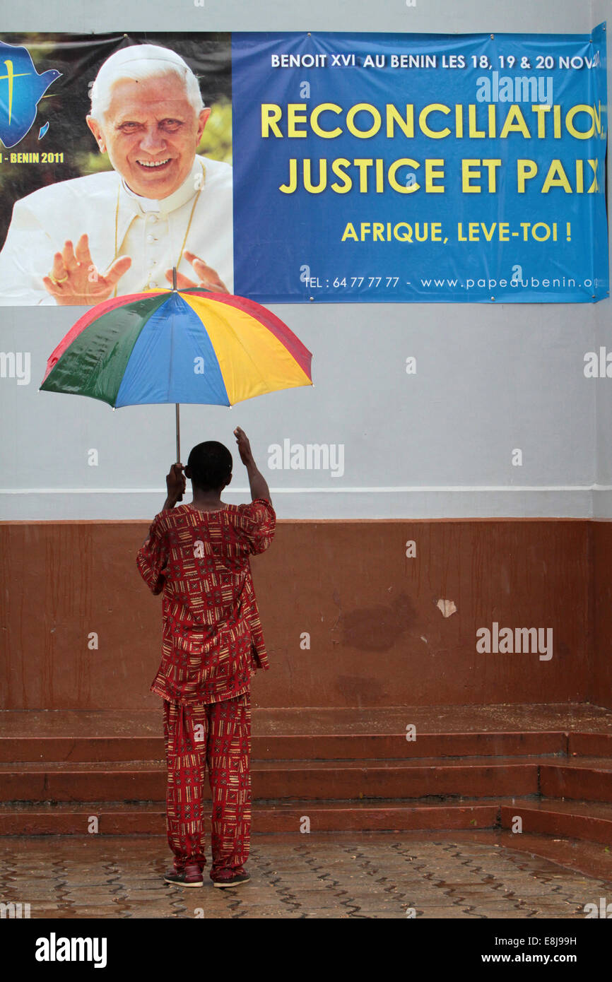 Africa, retro, portando un ombrello davanti a un poster che annuncia la visita di Papa Benedetto XVI in Benin. Foto Stock