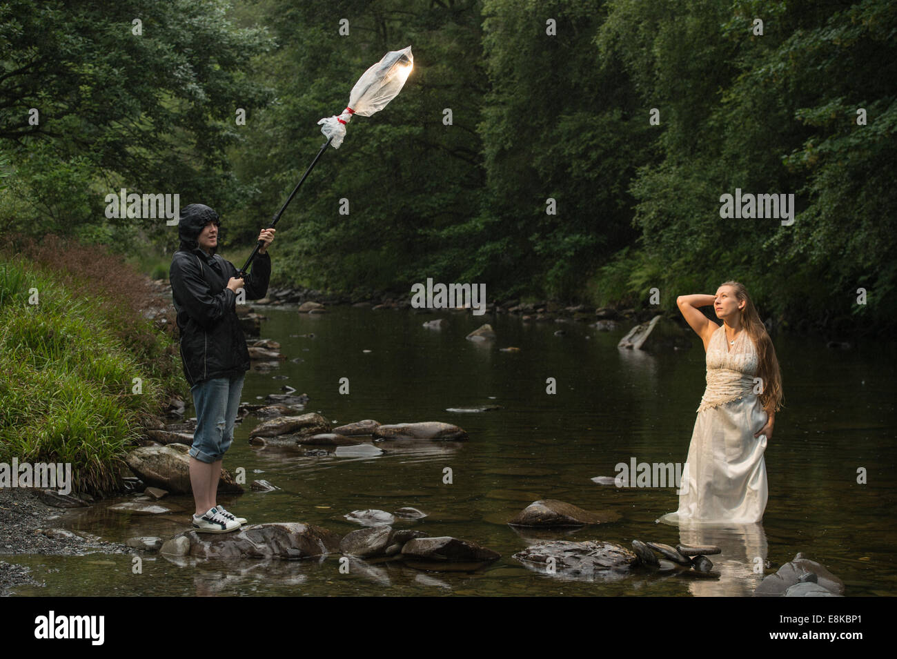Cestino il vestito photo shoot : un "sposa' che indossa il suo abito da sposa in piedi nel fiume - assistente di fotografi trattiene il braccio della luce Foto Stock